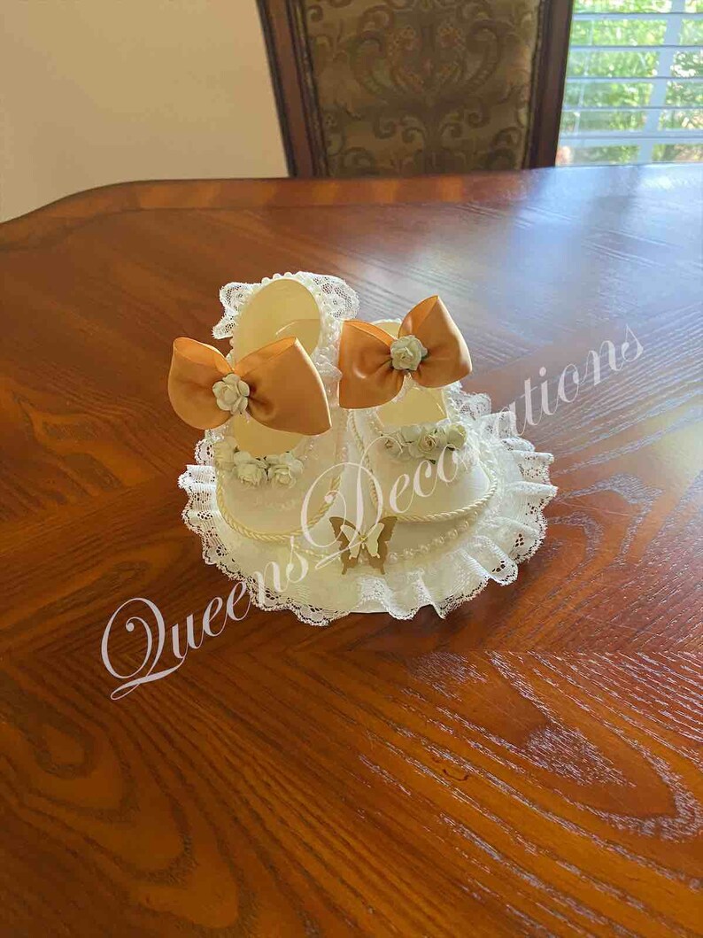 May include: Two white baby shoes with gold bows and lace trim. The shoes are on a white lace base with a gold butterfly.