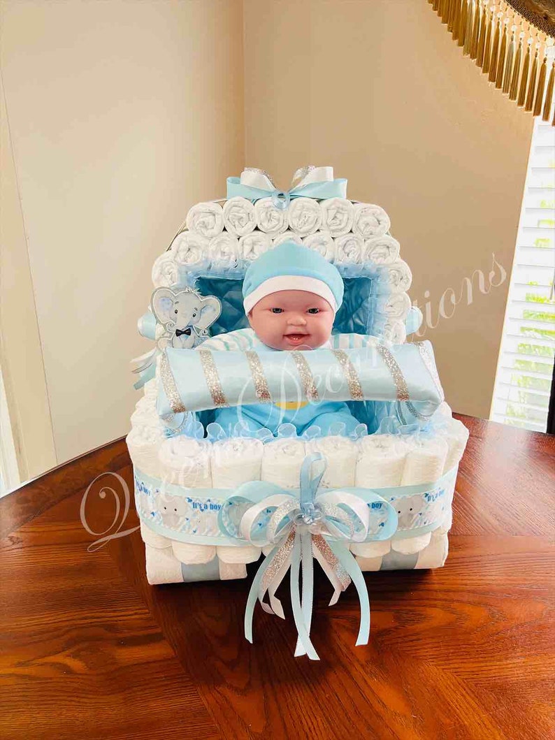 May include: A blue and white diaper cake shaped like a baby carriage. The cake is decorated with a blue ribbon and a silver bow. A baby doll is sitting in the carriage.