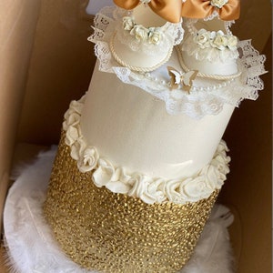 May include: A two-tiered white cake decorated with white icing and gold sequins. The top tier is adorned with a pair of white baby shoes with lace and gold accents, and a white butterfly.