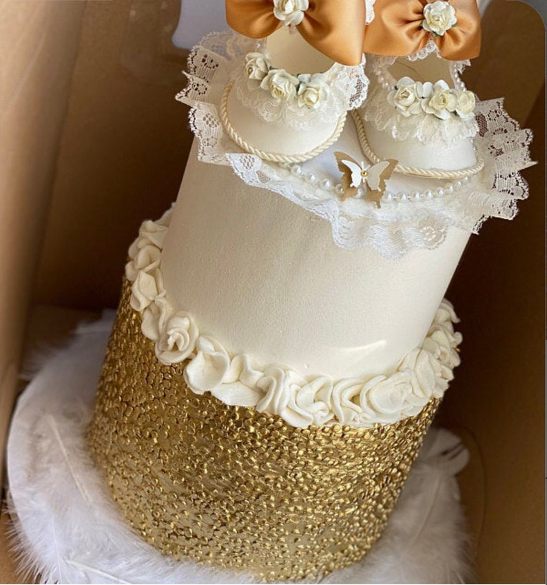 May include: A two-tiered white cake decorated with white icing and gold sequins. The top tier is adorned with a pair of white baby shoes with lace and gold accents, and a white butterfly.