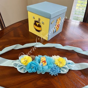 May include: A blue and yellow Winnie the Pooh themed baby shower sash with a Winnie the Pooh character holding a honey pot. The sash is decorated with blue and yellow flowers and a blue ribbon.