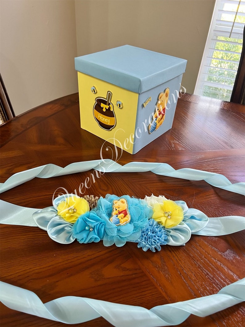 May include: A blue and yellow Winnie the Pooh themed baby shower sash with a Winnie the Pooh character holding a honey pot. The sash is decorated with blue and yellow flowers and a blue ribbon.