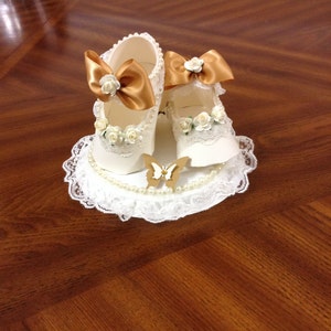 May include: A white lace doily with a pearl trim and a pair of white baby shoes with gold bows and white flowers. A gold butterfly is in the centre of the doily.