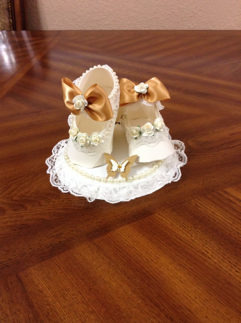 May include: A white lace doily with a pearl trim and a pair of white baby shoes with gold bows and white flowers. A gold butterfly is in the centre of the doily.