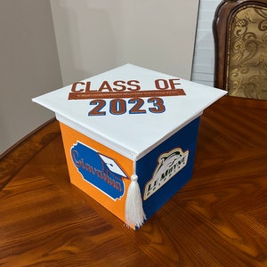 K&ouml;nnte beinhalten: Eine wei&szlig;e Abschluss-Hut-Box, die mit orangefarbenem und blauem Papier verziert ist. Die Oberseite der Box tr&auml;gt die Aufschrift "Class of 2023" in braunen Buchstaben. Die Seiten der Box tragen die Aufschrift "Congratulations" und "Le Moyne Dolphins" in orangefarbenen und blauen Buchstaben.