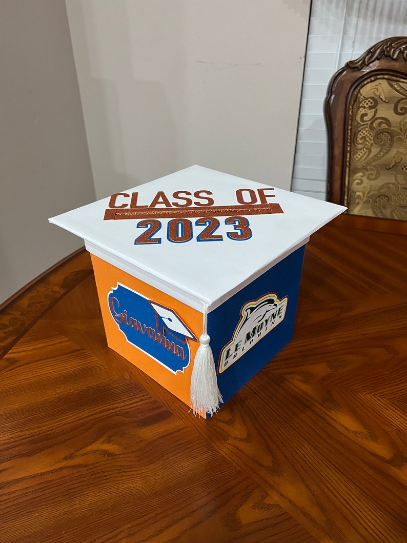 K&ouml;nnte beinhalten: Eine wei&szlig;e Abschluss-Hut-Box, die mit orangefarbenem und blauem Papier verziert ist. Die Oberseite der Box tr&auml;gt die Aufschrift "Class of 2023" in braunen Buchstaben. Die Seiten der Box tragen die Aufschrift "Congratulations" und "Le Moyne Dolphins" in orangefarbenen und blauen Buchstaben.