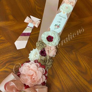 May include: A pink and gold sash with a large bow and a floral arrangement. The sash has the text "Mommy To Be" on a tag. The floral arrangement includes pink, white, burgundy, and gold flowers.