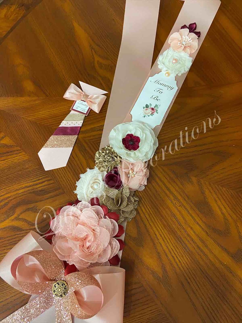 May include: A pink and gold sash with a large bow and a floral arrangement. The sash has the text "Mommy To Be" on a tag. The floral arrangement includes pink, white, burgundy, and gold flowers.