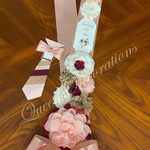 May include: A pink and gold sash with a large bow and a tag that says "Mommy To Be". The sash is decorated with several fabric flowers in shades of pink, white, burgundy, and gold.