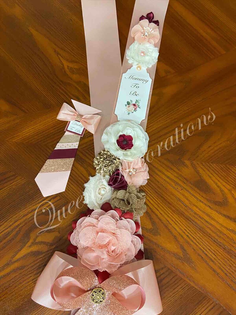 May include: A pink and gold sash with a large bow and a tag that says "Mommy To Be". The sash is decorated with several fabric flowers in shades of pink, white, burgundy, and gold.