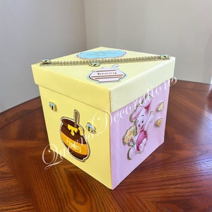 May include: A yellow and pink gift box with a Winnie the Pooh character on the side. The box has a gold chain and a honey pot with a bee on it. The text on the box says "Winnie's Baby Shower".
