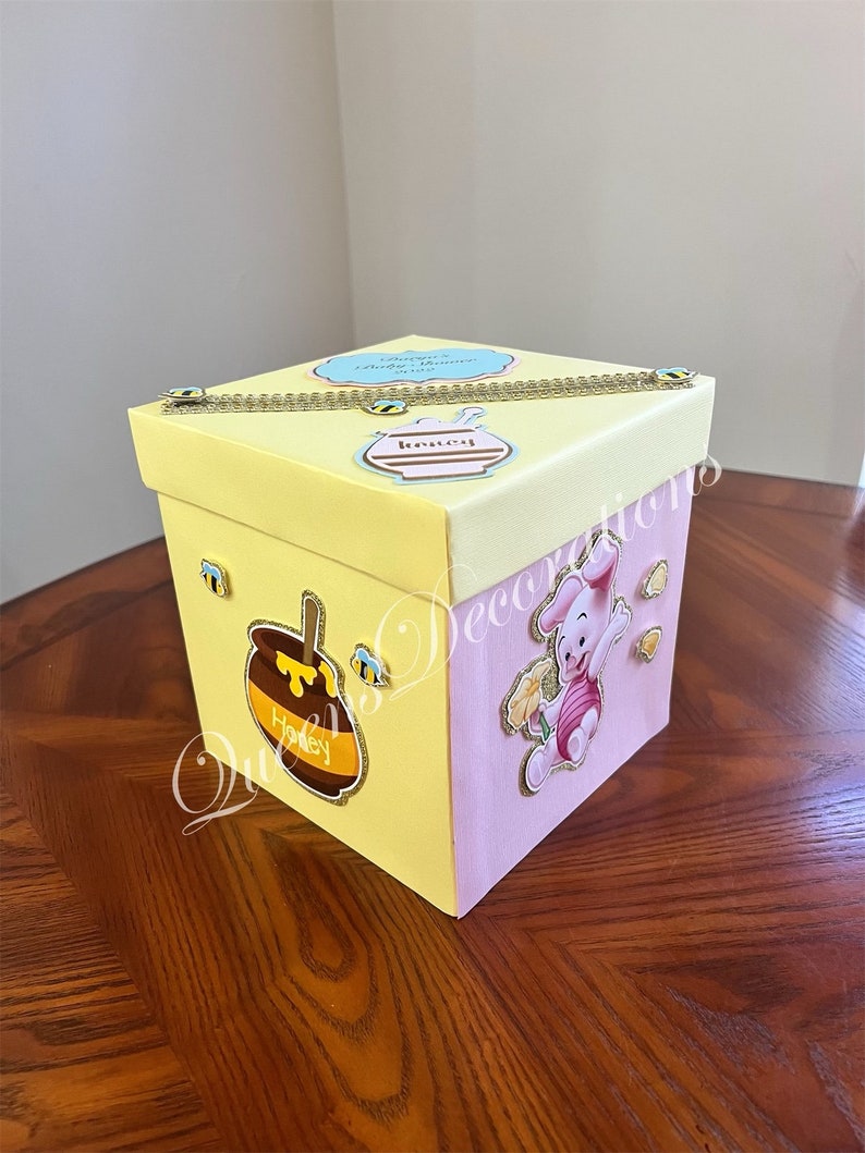 May include: A yellow and pink gift box with a Winnie the Pooh character on the side. The box has a gold chain and a honey pot with a bee on it. The text on the box says "Winnie's Baby Shower".