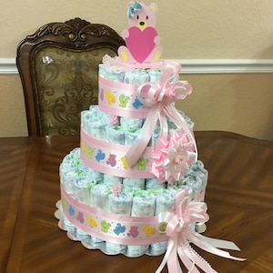 May include: A three-tiered nappy cake decorated with pink and white ribbons, a pink bear topper, and colourful bear print ribbon. The cake is on a white scalloped base.