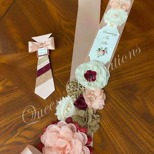 May include: A pink and gold sash with a bow and flowers, a tag that says "Mommy To Be", and a matching tie with the text "Daddy To Be".