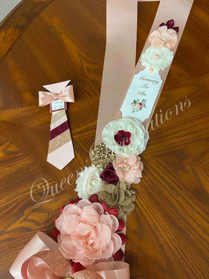 May include: A pink and gold sash with a bow and flowers, a tag that says "Mommy To Be", and a matching tie with the text "Daddy To Be".