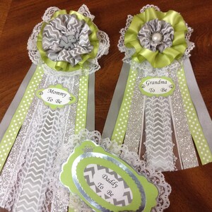 May include: Three green and silver sashes with white lace trim and a gray and white chevron pattern. The sashes have a silver glitter ribbon and a green tag with the text "Mommy To Be", "Daddy To Be", and "Grandma To Be".