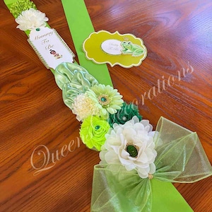 May include: Green and white sash with a bow, decorated with flowers and a frog prince charm. The sash has a tag that says "Mommy To Be" and another tag that says "Daddy To Be".