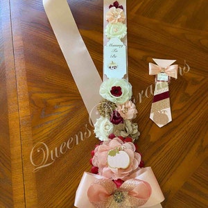 May include: A pink and gold satin sash with a bow and a pumpkin-themed flower arrangement. The sash has the text "Mommy To Be" on a white tag. A matching tie with a pumpkin design is also included.