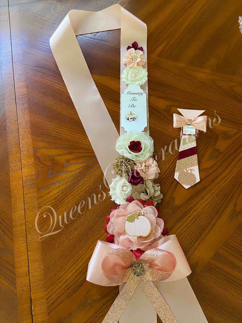 May include: A pink and gold satin sash with a bow and a pumpkin-themed flower arrangement. The sash has the text "Mommy To Be" on a white tag. A matching tie with a pumpkin design is also included.