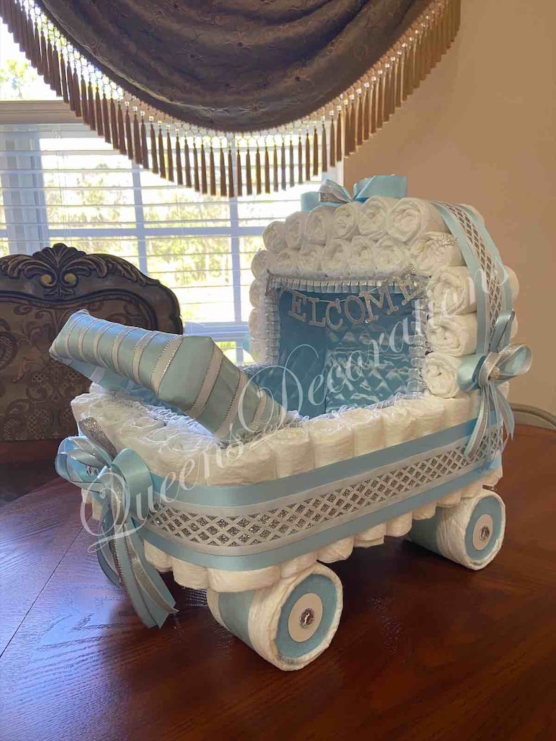 Puede incluir: Un pastel de pa&ntilde;ales azul y blanco con forma de cochecito de beb&eacute;. El cochecito est&aacute; decorado con una cinta plateada y un letrero que dice "Bienvenido".