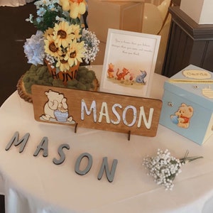 May include: A Winnie the Pooh themed baby shower table setting with a blue and white balloon arch, a Winnie the Pooh figurine, a floral arrangement, a wooden sign with the name "MASON" and a blue gift box with a Winnie the Pooh design. The table is covered with a white tablecloth.