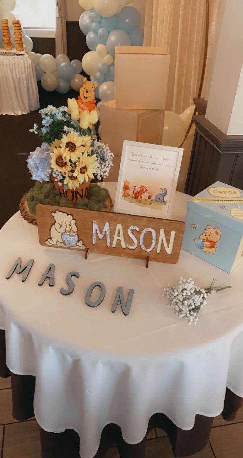 May include: A Winnie the Pooh themed baby shower table setting with a blue and white balloon arch, a Winnie the Pooh figurine, a floral arrangement, a wooden sign with the name "MASON" and a blue gift box with a Winnie the Pooh design. The table is covered with a white tablecloth.