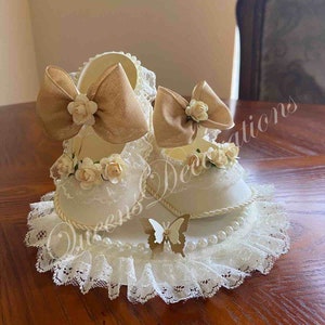 May include: A pair of white baby shoes with lace trim and brown bows, decorated with white flowers and pearls, on a white lace doily with a gold butterfly.