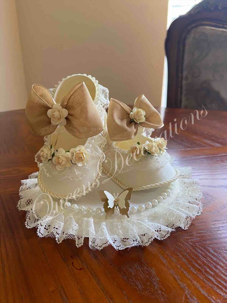 May include: A pair of white baby shoes with lace trim and brown bows, decorated with white flowers and pearls, on a white lace doily with a gold butterfly.