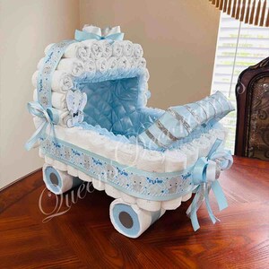 May include: A blue and white diaper cake shaped like a baby carriage. The carriage is decorated with blue and white ribbon, a silver ribbon, and a small stuffed elephant. The cake is sitting on a wooden table.