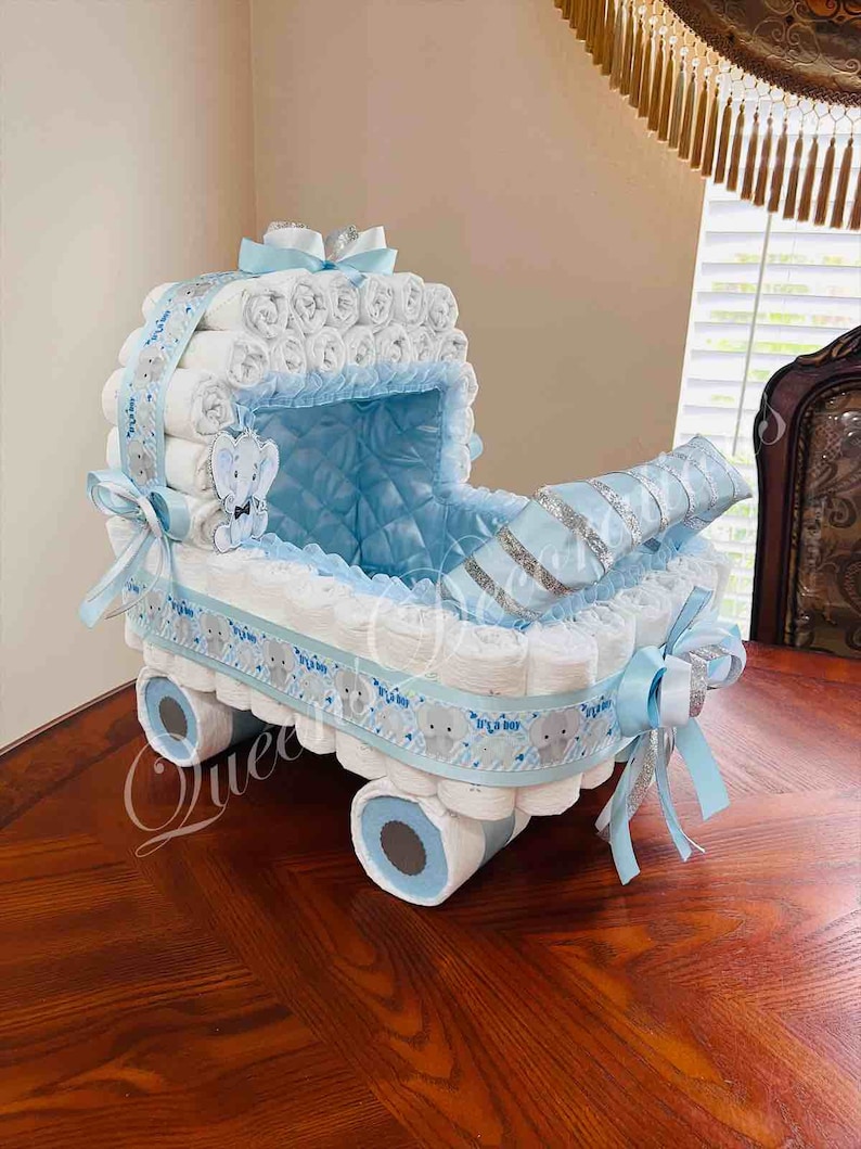 May include: A blue and white diaper cake shaped like a baby carriage. The carriage is decorated with blue and white ribbon, a silver ribbon, and a small stuffed elephant. The cake is sitting on a wooden table.