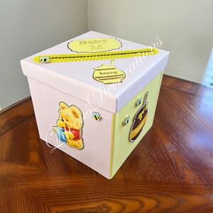 May include: A pink and yellow gift box with a Winnie the Pooh character on the side. The box has a yellow ribbon and a tag that says "Baby M".