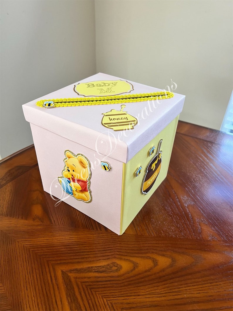 May include: A pink and yellow gift box with a Winnie the Pooh character on the side. The box has a yellow ribbon and a tag that says "Baby M".