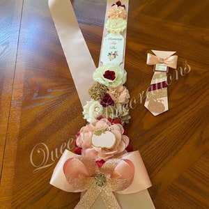 May include: A pink and gold sash with a bow and a white tag that says "Mommy To Be". The sash is decorated with burgundy, peach, and gold flowers. A matching burgundy and gold tie is also included.