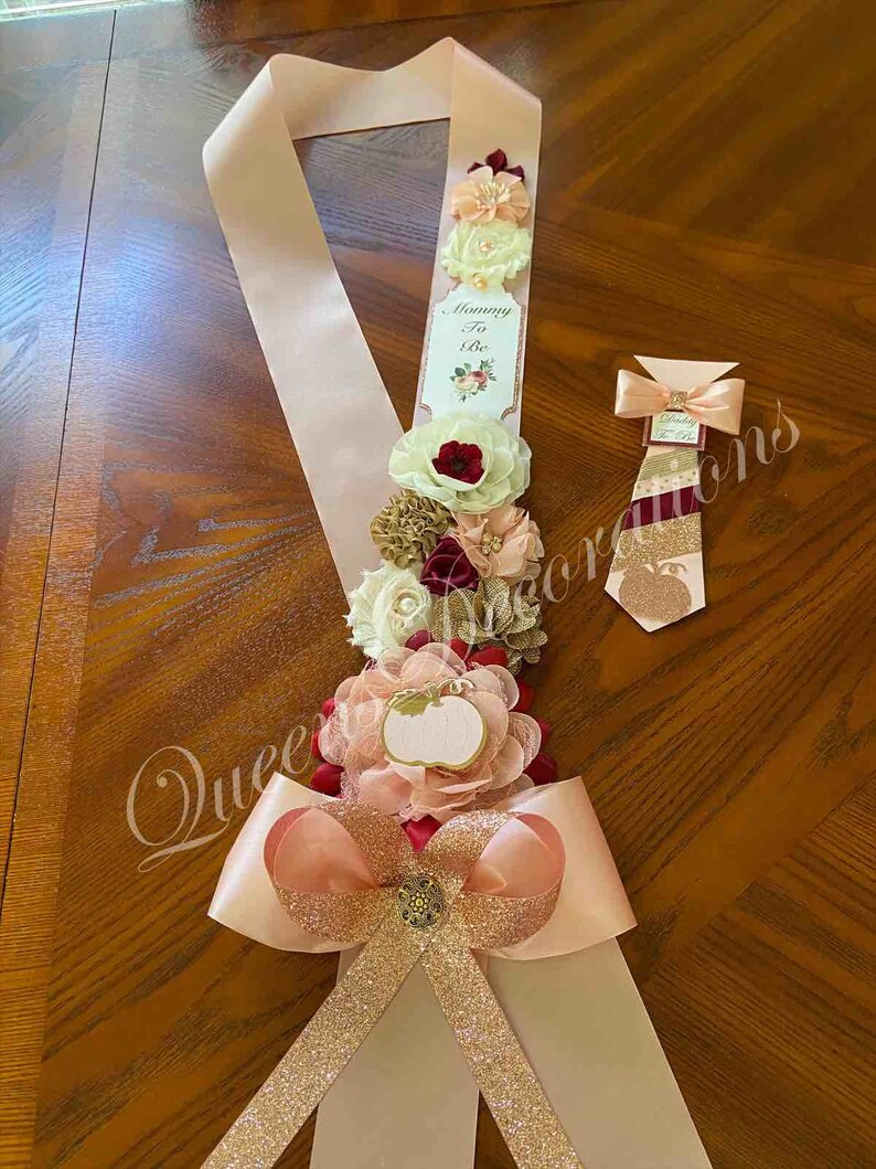 May include: A pink and gold sash with a bow and a white tag that says "Mommy To Be". The sash is decorated with burgundy, peach, and gold flowers. A matching burgundy and gold tie is also included.