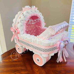 Puede incluir: Un pastel de pa&ntilde;ales rosa y blanco con forma de cochecito. El cochecito est&aacute; decorado con cinta plateada, un lazo rosa y un ribete de encaje blanco. El cochecito tiene un forro rosa y un letrero que dice "Bienvenido".