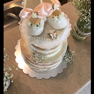 May include: A two-tiered cake decorated with white lace, pearl beads, and a pair of white baby shoes with pink bows. The shoes are topped with white flowers and a gold butterfly.