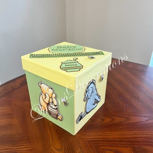 May include: A yellow and green gift box with a Winnie the Pooh and Eeyore theme. The box has a gold ribbon and the text "Baby Martinez" on the top. The box is decorated with a honey pot, bees, and the characters Winnie the Pooh and Eeyore.