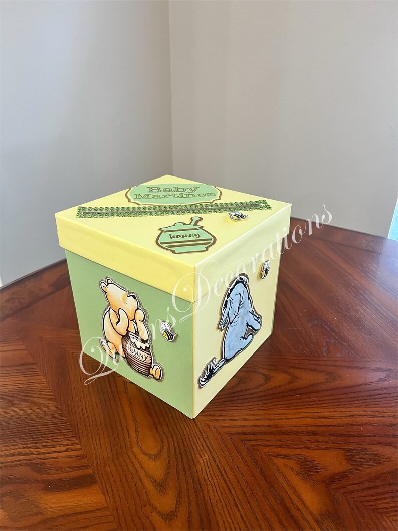 May include: A yellow and green gift box with a Winnie the Pooh and Eeyore theme. The box has a gold ribbon and the text "Baby Martinez" on the top. The box is decorated with a honey pot, bees, and the characters Winnie the Pooh and Eeyore.