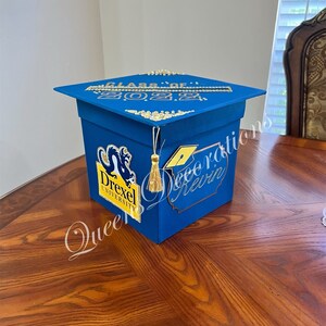 K&ouml;nnte beinhalten: Blaue Box in Form einer Abschluss-Kappe mit goldenen Akzenten. Die Box hat ein Drexel University Logo und den Text "Class of 2022".