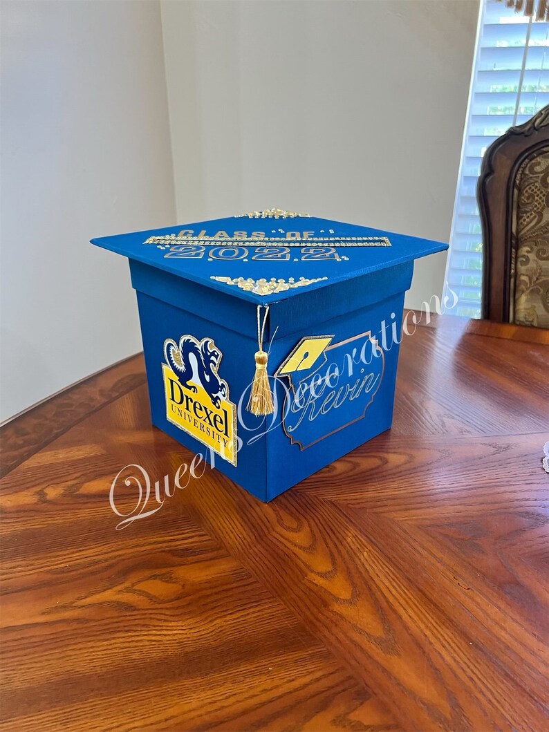 K&ouml;nnte beinhalten: Blaue Box in Form einer Abschluss-Kappe mit goldenen Akzenten. Die Box hat ein Drexel University Logo und den Text "Class of 2022".