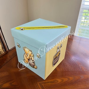 May include: A light blue and yellow gift box with a Winnie the Pooh character and a honey pot design. The box has a yellow ribbon and a bee design.