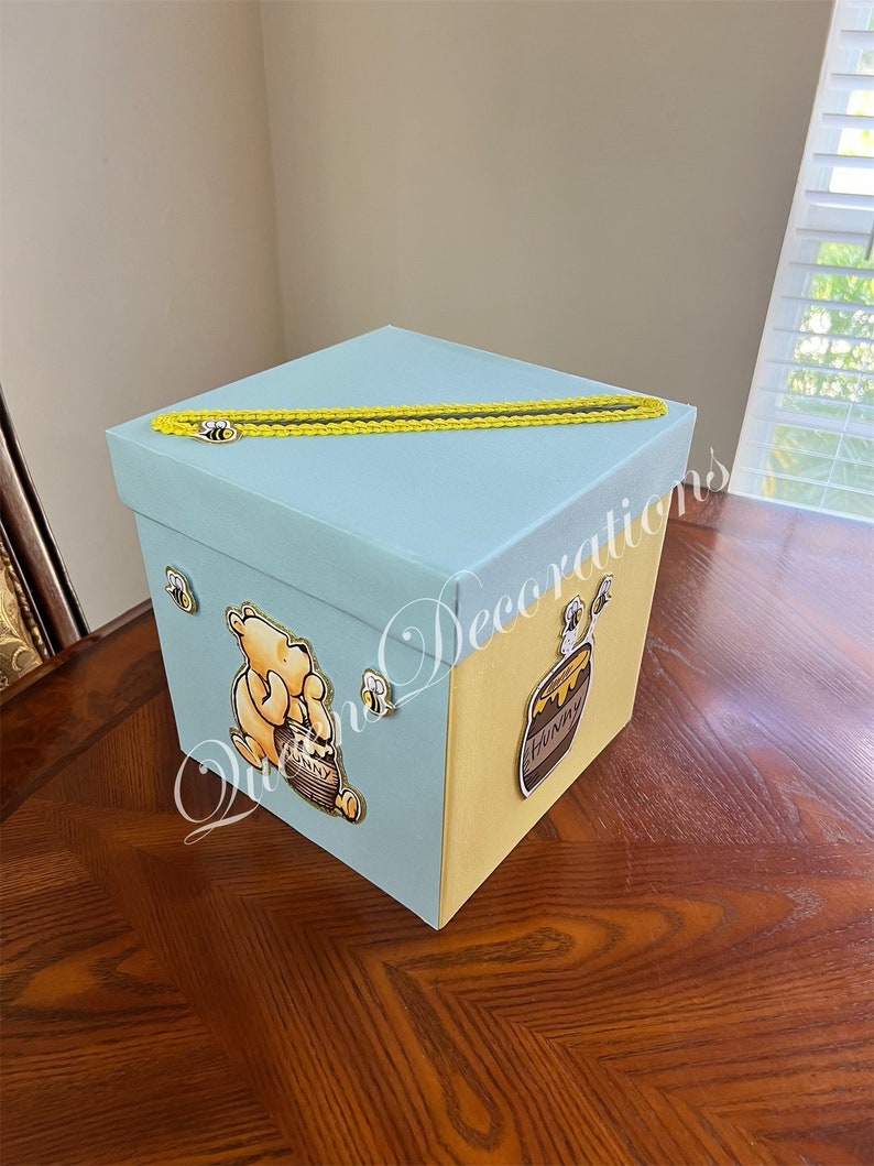 May include: A light blue and yellow gift box with a Winnie the Pooh character and a honey pot design. The box has a yellow ribbon and a bee design.