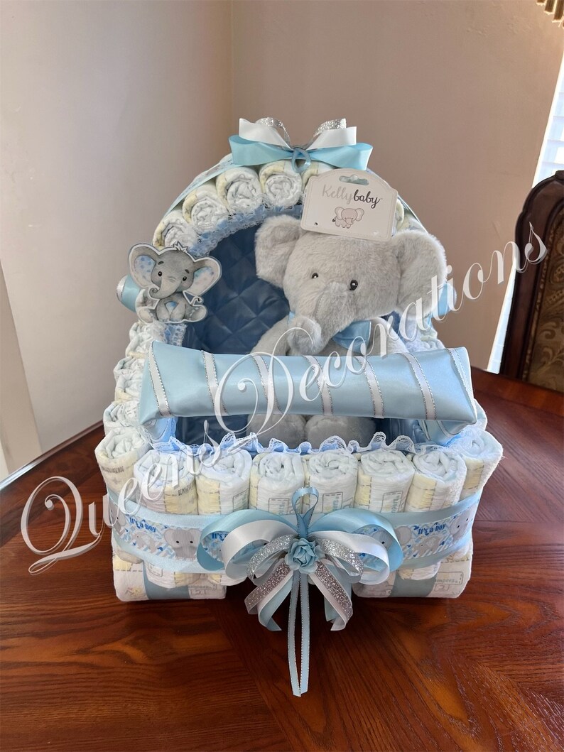 May include: A blue and white diaper cake shaped like a baby carriage with a gray elephant plush toy sitting inside. The cake is decorated with a blue and white ribbon and a small white tag that says "Kelly baby".