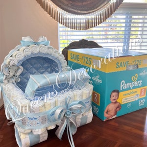 May include: A blue and white diaper cake shaped like a baby stroller with a gray elephant on top. The stroller is decorated with a white ribbon and a silver bow. The cake is sitting on a wooden table next to a box of Pampers swaddlers diapers. The box says "Save up to $7.25" and "180 diapers".