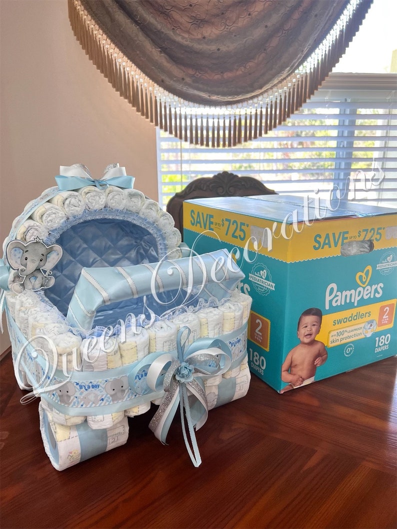 May include: A blue and white diaper cake shaped like a baby stroller with a gray elephant on top. The stroller is decorated with a white ribbon and a silver bow. The cake is sitting on a wooden table next to a box of Pampers swaddlers diapers. The box says "Save up to $7.25" and "180 diapers".
