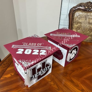 K&ouml;nnte beinhalten: Zwei Abschluss-Hut-Boxen, verziert mit Strasssteinen und dem Text "Klasse von 2022". Eine Box ist dunkelrot mit einem wei&szlig;en Jesuiten-Logo und die andere ist wei&szlig; mit einem Pfotenabdruck.