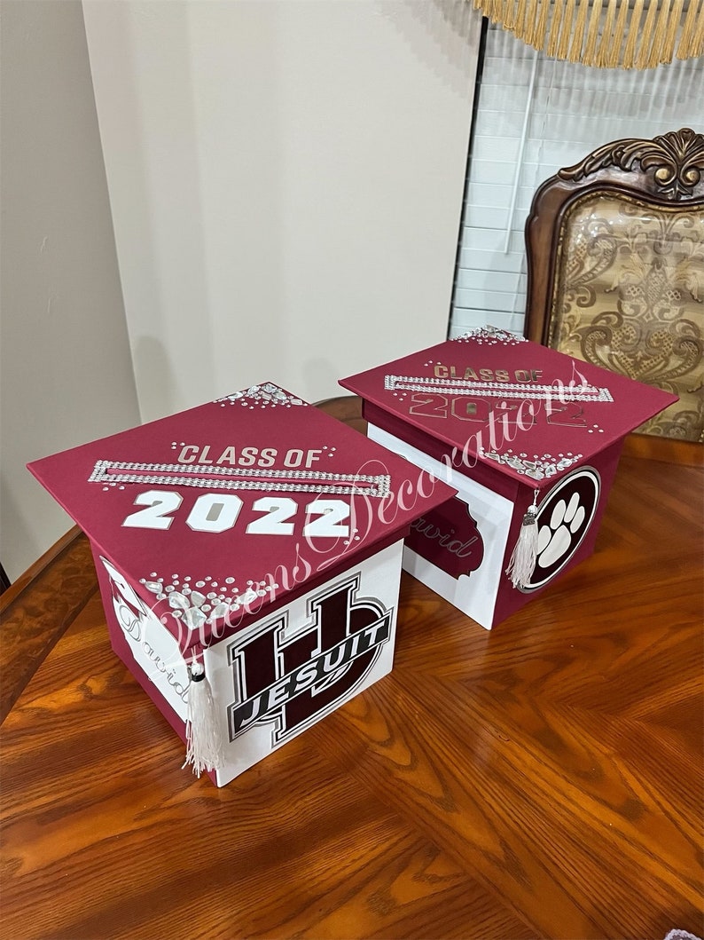 K&ouml;nnte beinhalten: Zwei Abschluss-Hut-Boxen, verziert mit Strasssteinen und dem Text "Klasse von 2022". Eine Box ist dunkelrot mit einem wei&szlig;en Jesuiten-Logo und die andere ist wei&szlig; mit einem Pfotenabdruck.