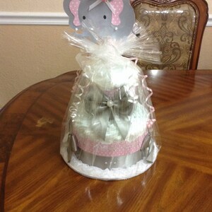 May include: A three-tiered diaper cake decorated with pink and white polka dots, a silver bow, and a gray elephant topper with pink accents. The cake is wrapped in clear cellophane and sits on a wooden table.