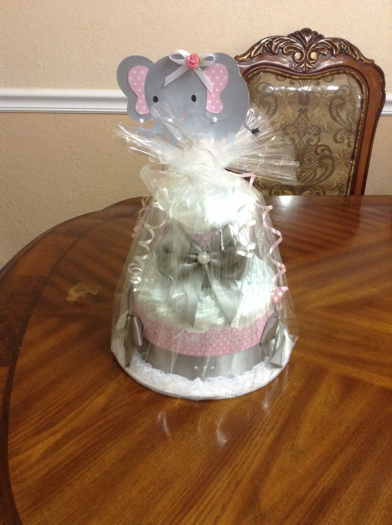 May include: A three-tiered diaper cake decorated with pink and white polka dots, a silver bow, and a gray elephant topper with pink accents. The cake is wrapped in clear cellophane and sits on a wooden table.
