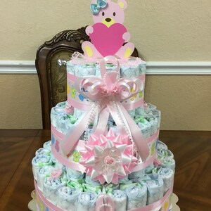 May include: A three-tiered nappy cake decorated with pink and white ribbons, a pink flower, and a pink bear holding a pink heart. The cake is made of rolled nappies and is a popular baby shower gift.